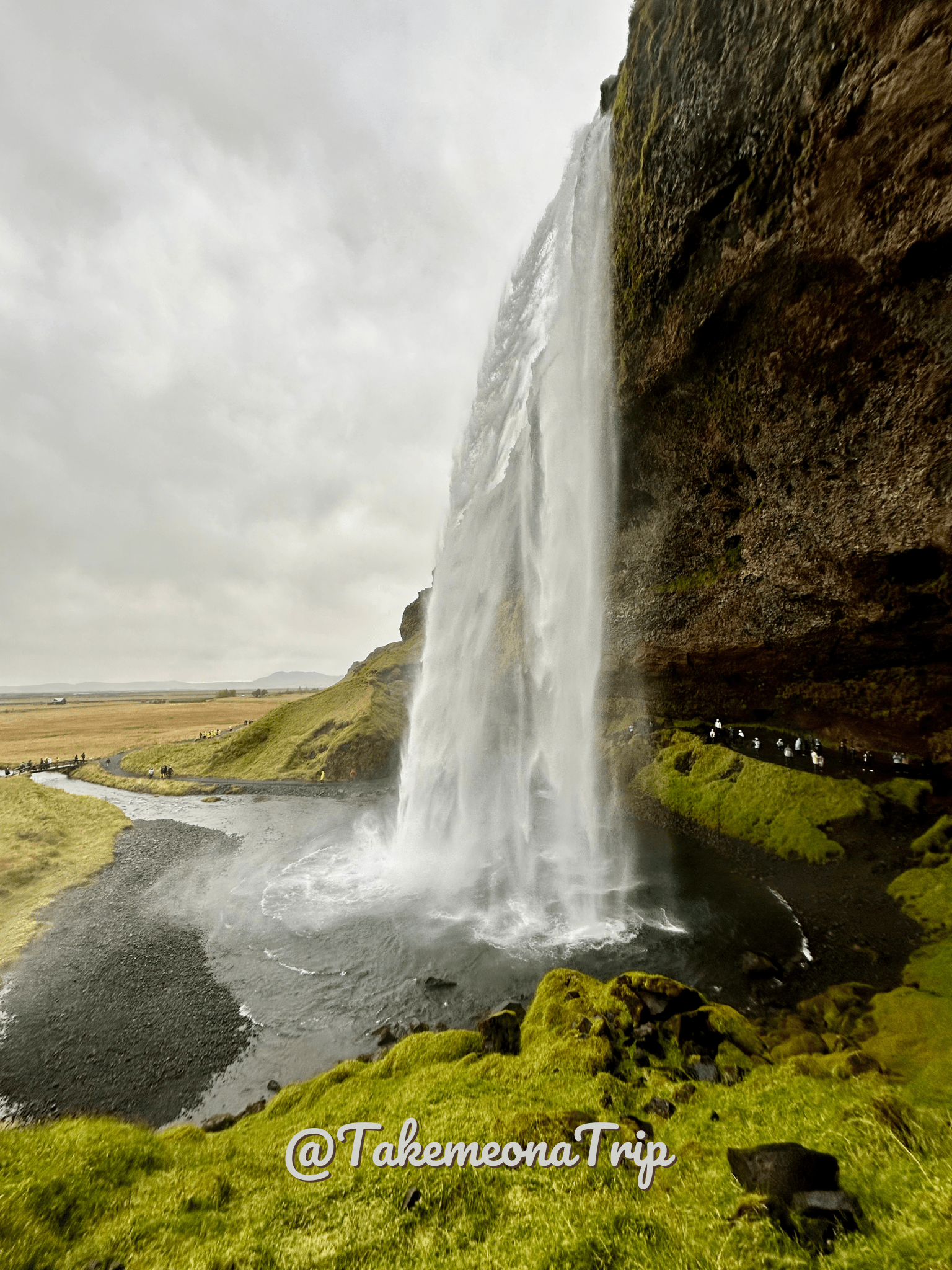 Cascata di Seljalandfoss vista di lato. Cielo grigio, muschio verde, cascata con persone dietro.